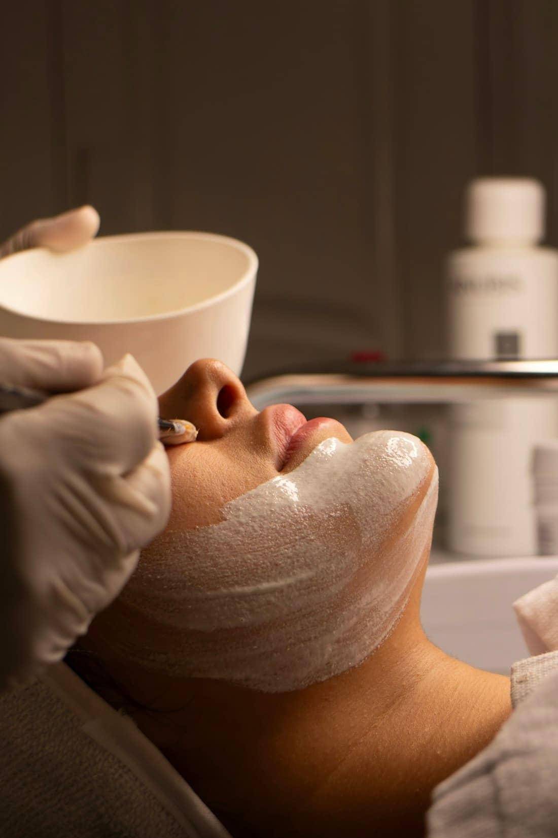 Gloved hands applying a clinical facial treatment from a mixing bowl in warm light